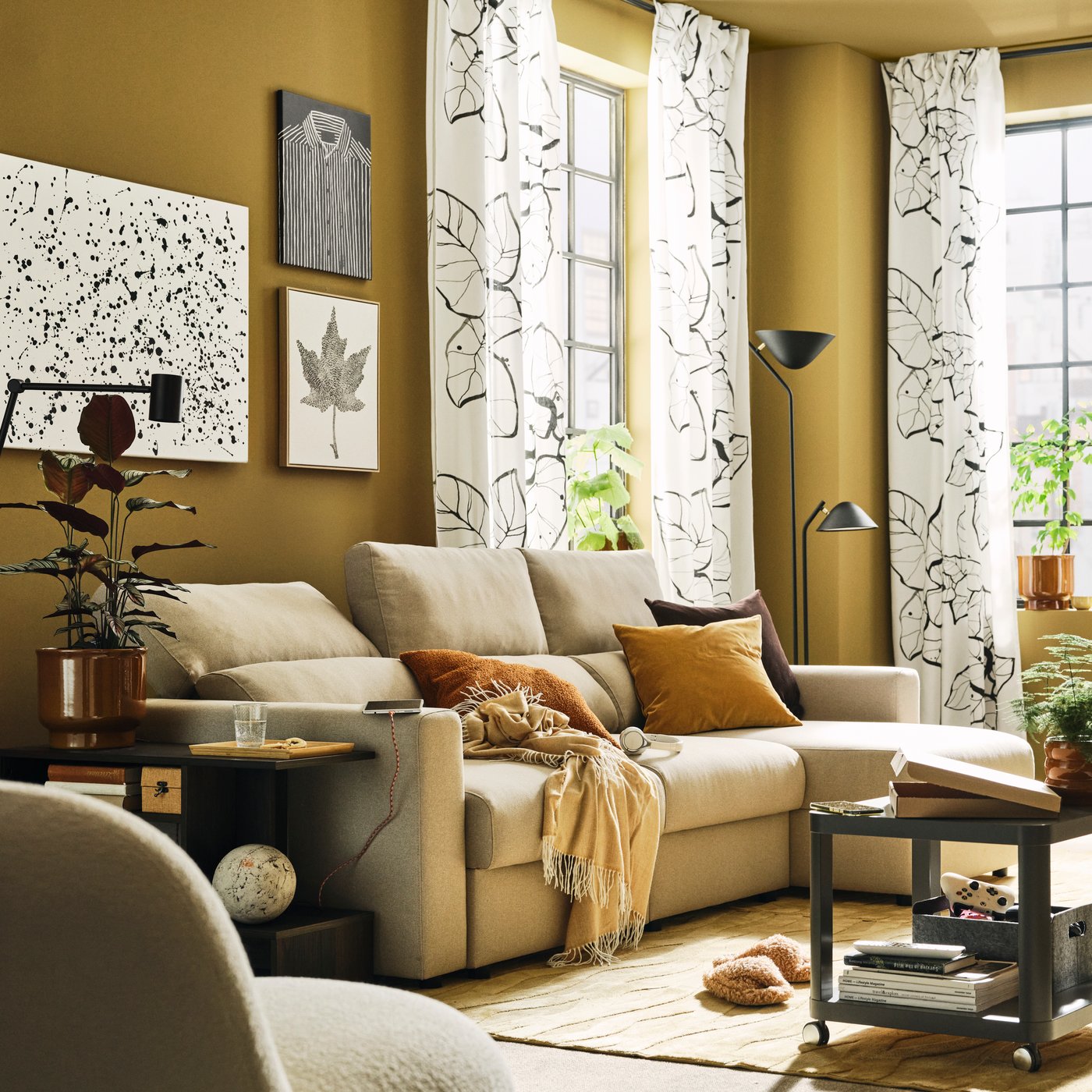 A light living room with a light-beige MALMKÖPING sofa with chaise longue flanked by side tables, a floor lamp and plants.