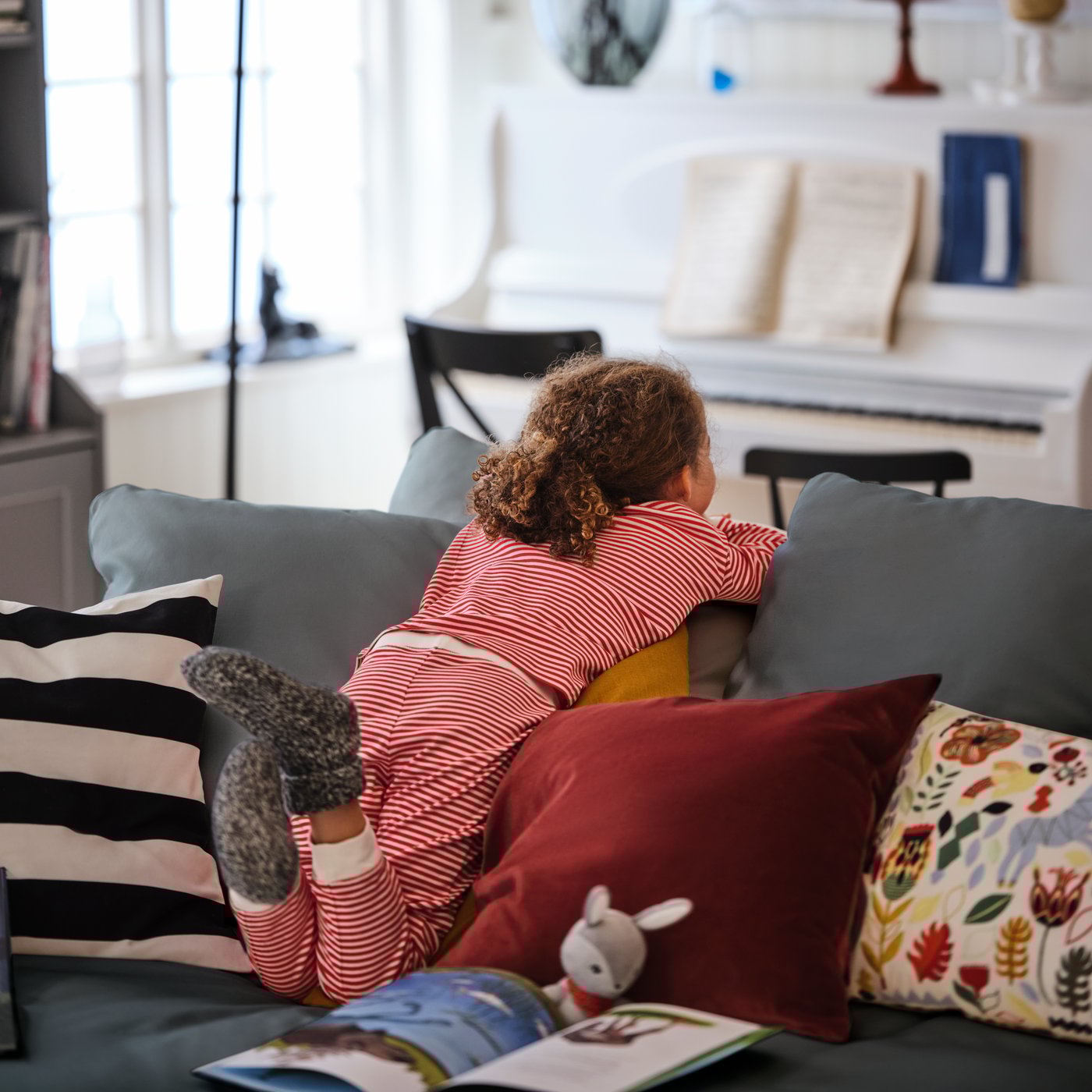 A young girl leans into the backrest of a grey GRÖNLID four-seat sofa with chaise longues, peeking over its back.