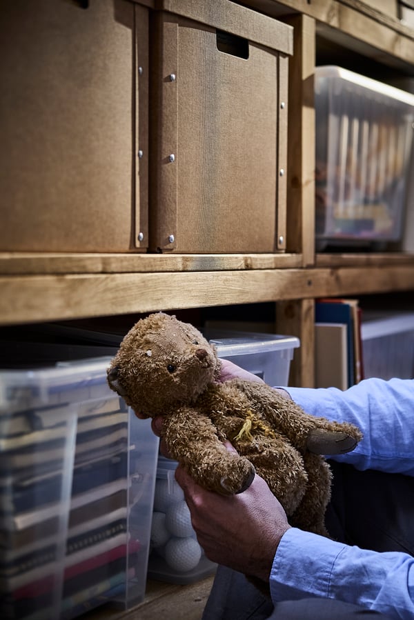 På en reol i en garage står fyldte SAMLA kasser med låg og foran kasserne holder to hænder en gammel bamse.