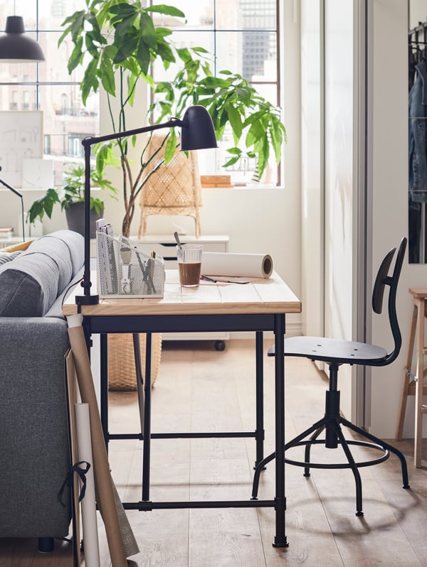 Uma secretária com tampo de madeira e pés em preto, um copo, um vaso de plantas e outros objetos, uma cadeira giratória e as costas de um sofá.