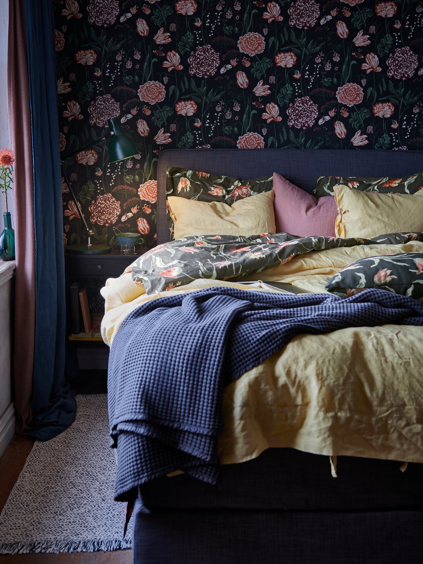 A bed made with yellow, blue and floral bed linen, and a blue folded throw, on a rug by a wall with dark floral pattern.