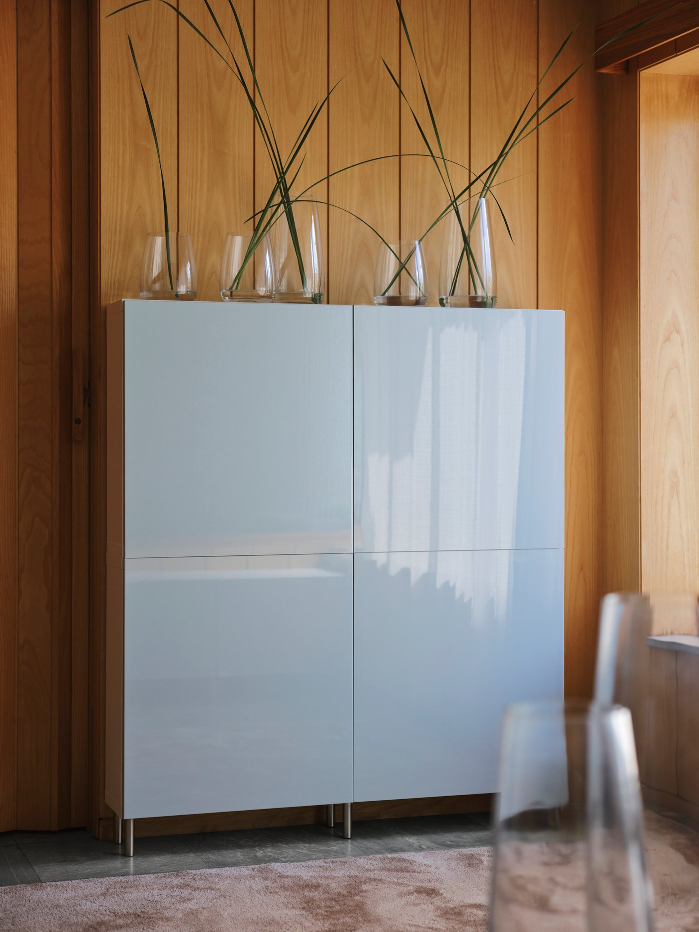 A highboard with four square doors with high-gloss fronts and several transparent vases containing branches on the top. 
