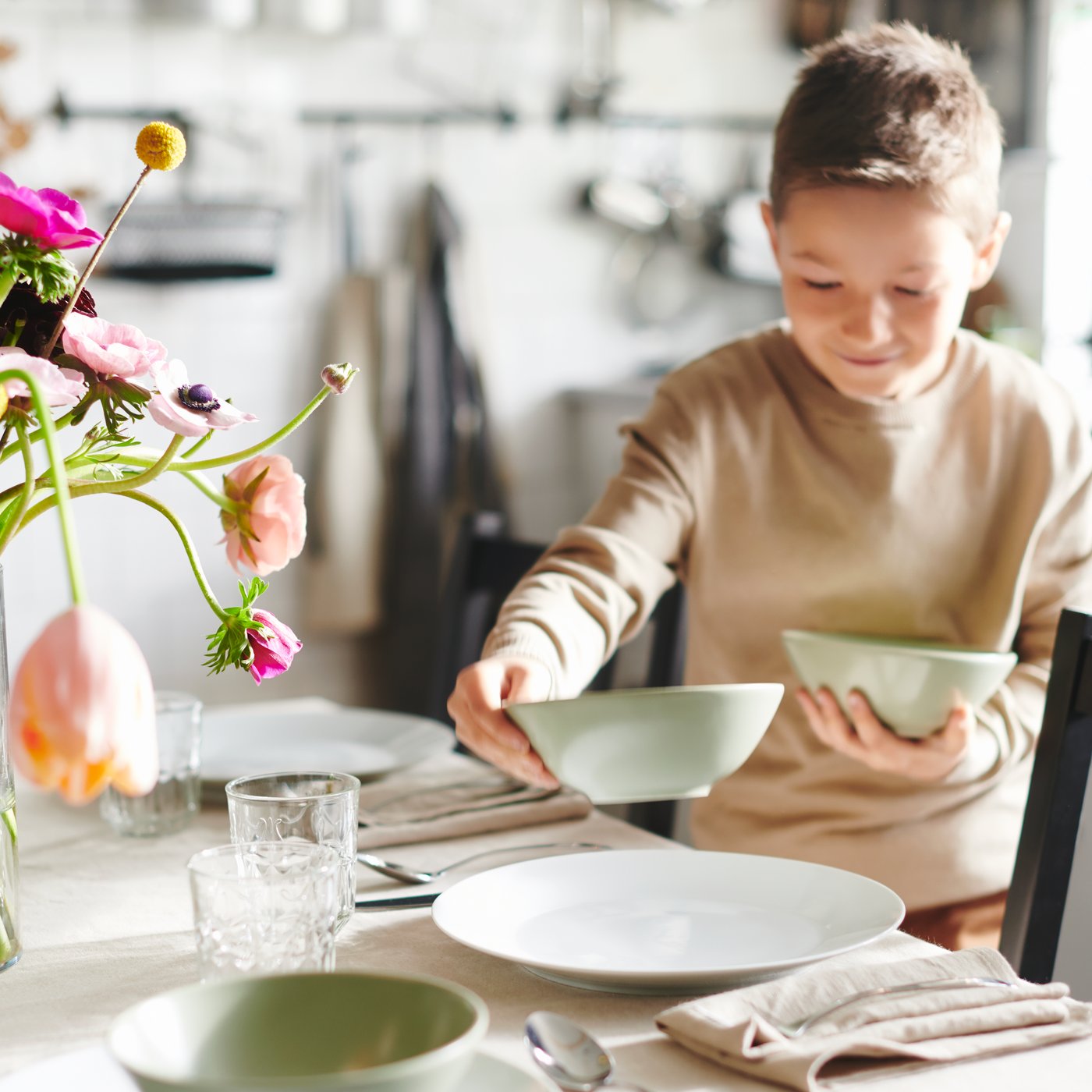Ein Kind deckt den Esstisch mit FÄRKLAR Schüsseln. Auf dem Tisch befindet sich ein Blumenstrauß.