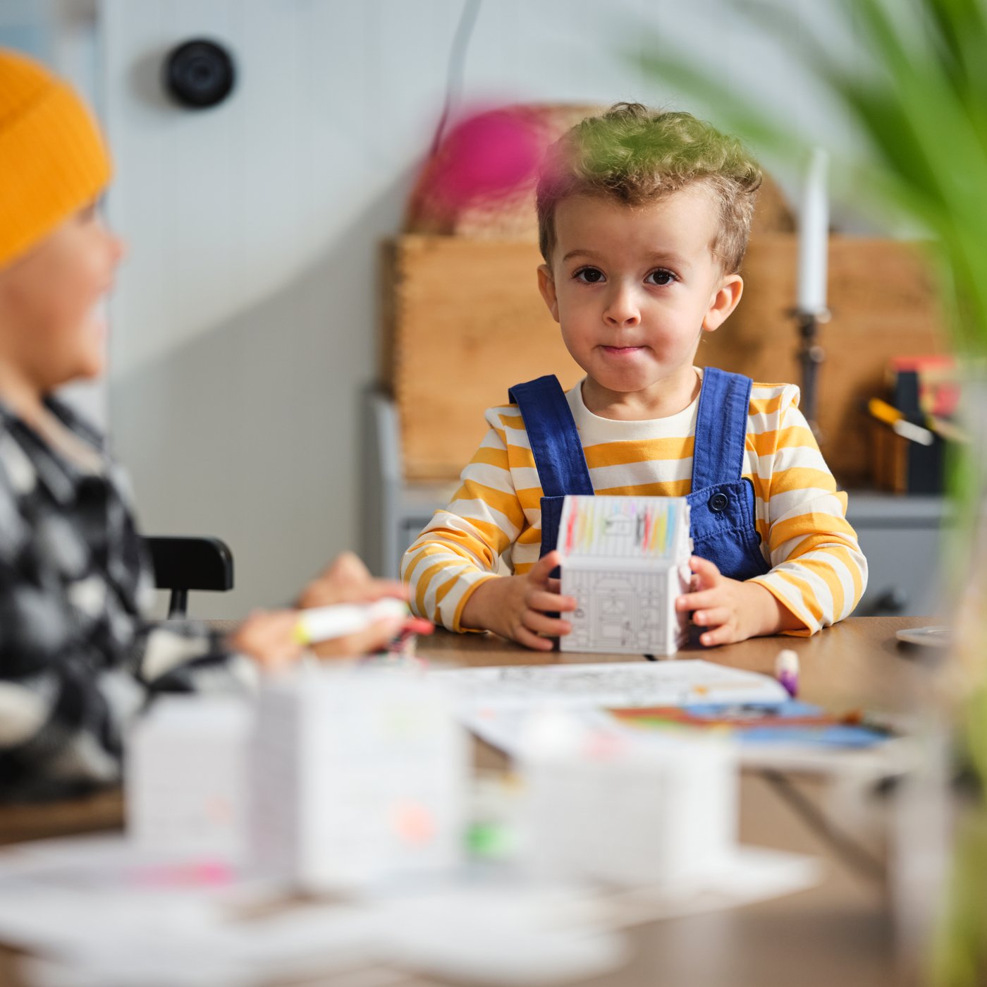 A young boy sitting at the end of a long dining table, holding a MÅLA cardboard town template, folded into a house shape.