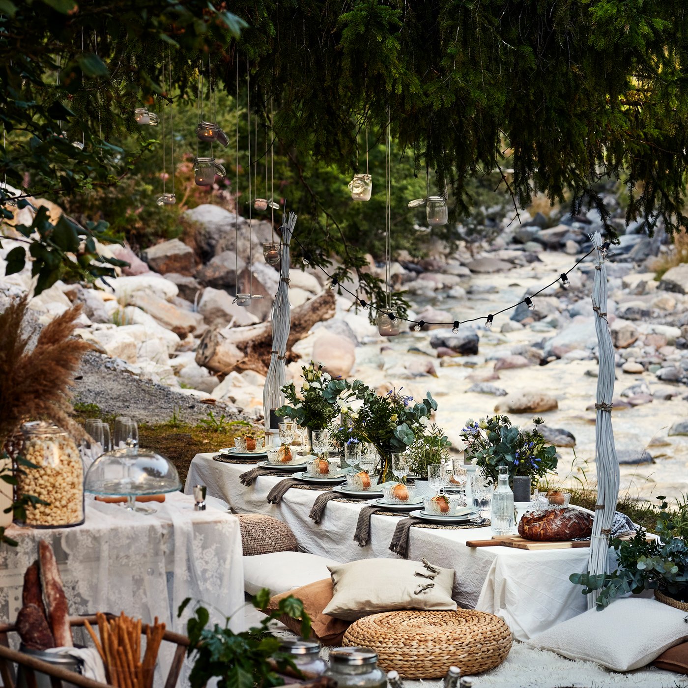 Eine lange Tafel mit Tischdecke, Geschirr und vielen Blumen steht in der natur neben einem Bach. Darum sind verschiedene Sitzkissen und Poufs angeordnet und darüber hängen Laternen und eine Lichterkette.