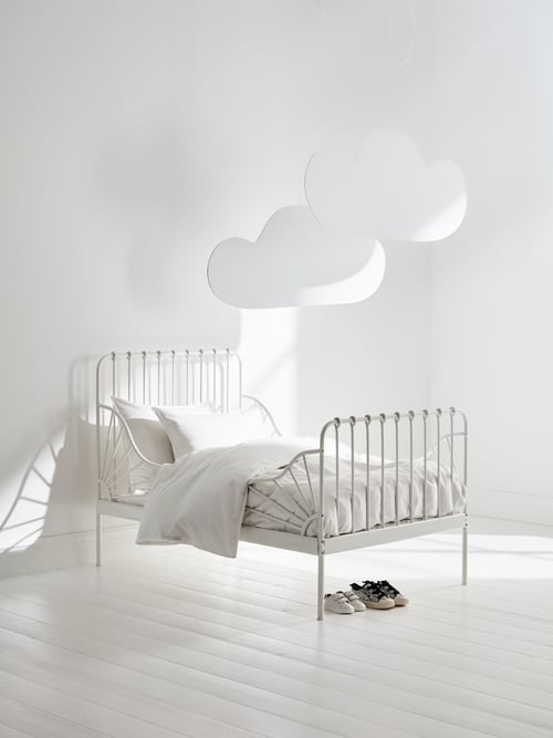 A MINNEN extendable bed frame in white has white bed linen on it, decorative clouds hanging above and kids' shoes beneath.