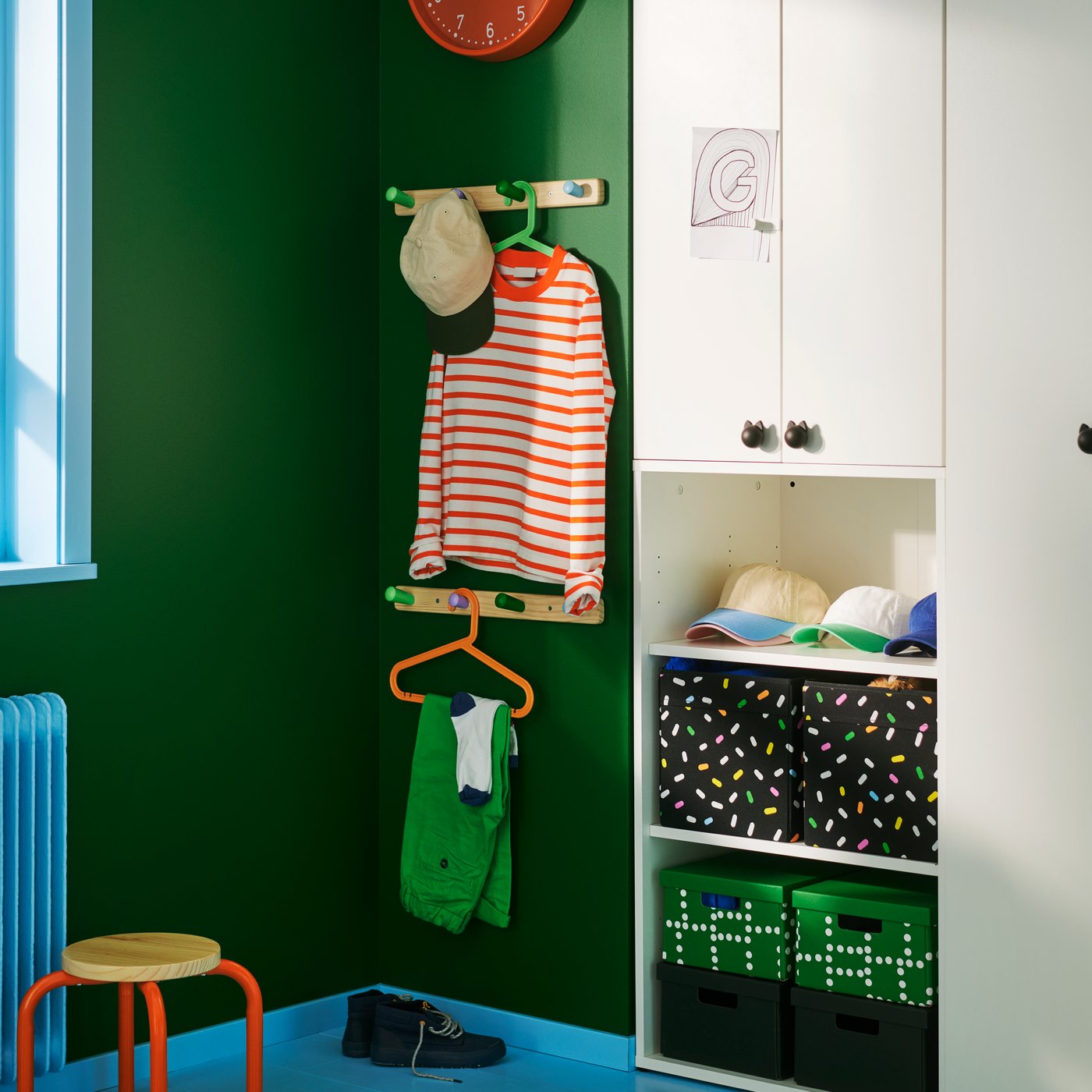 A BAGIS children’s coat-hanger in orange, on a wall rail in a green-walled children’s room with a wardrobe and storage boxes.