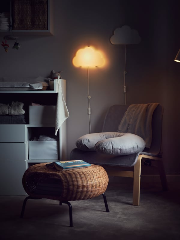 A dim room lit by a cloud shaped wall lamp, a grey armchair, a rattan GAMLEHULT footstool with a cushion resting on it.