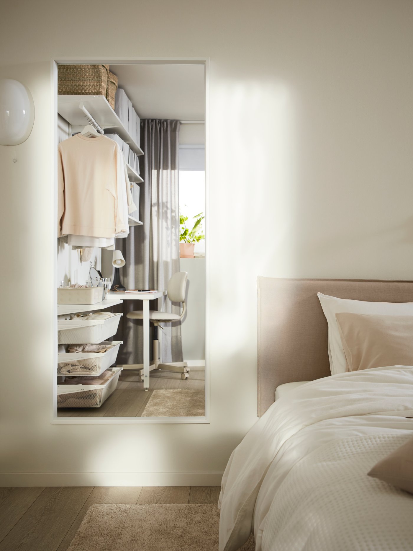 Un espejo NISSEDAL en una pared junto a una cama en el que se ve una ventana con cortinas de color gris claro y una cortina junto a un almacenaje de pared abierto.