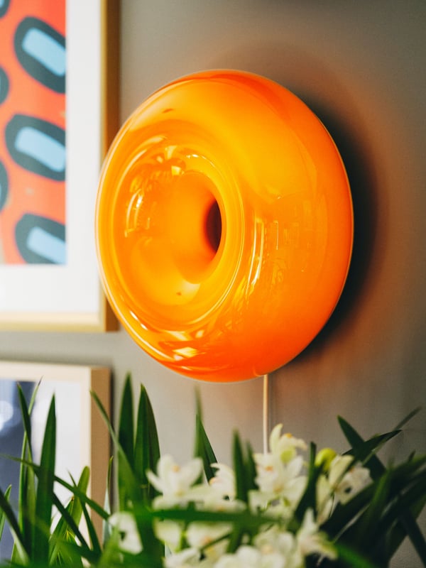 A round orange glass VARMBLIXT LED table/wall lamp and some framed posters are mounted on a wall near some white flowers.