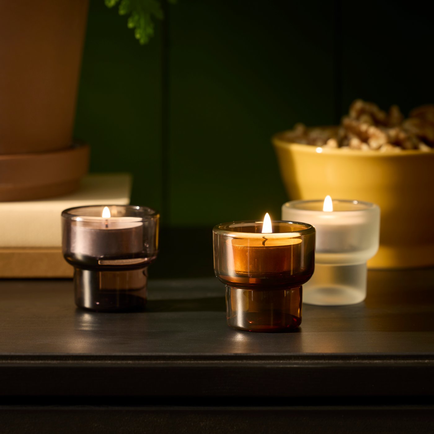 Three LADUSVALA tealight holders in mixed colours are arranged on a dark table, together with a plant and a decorative bowl.
