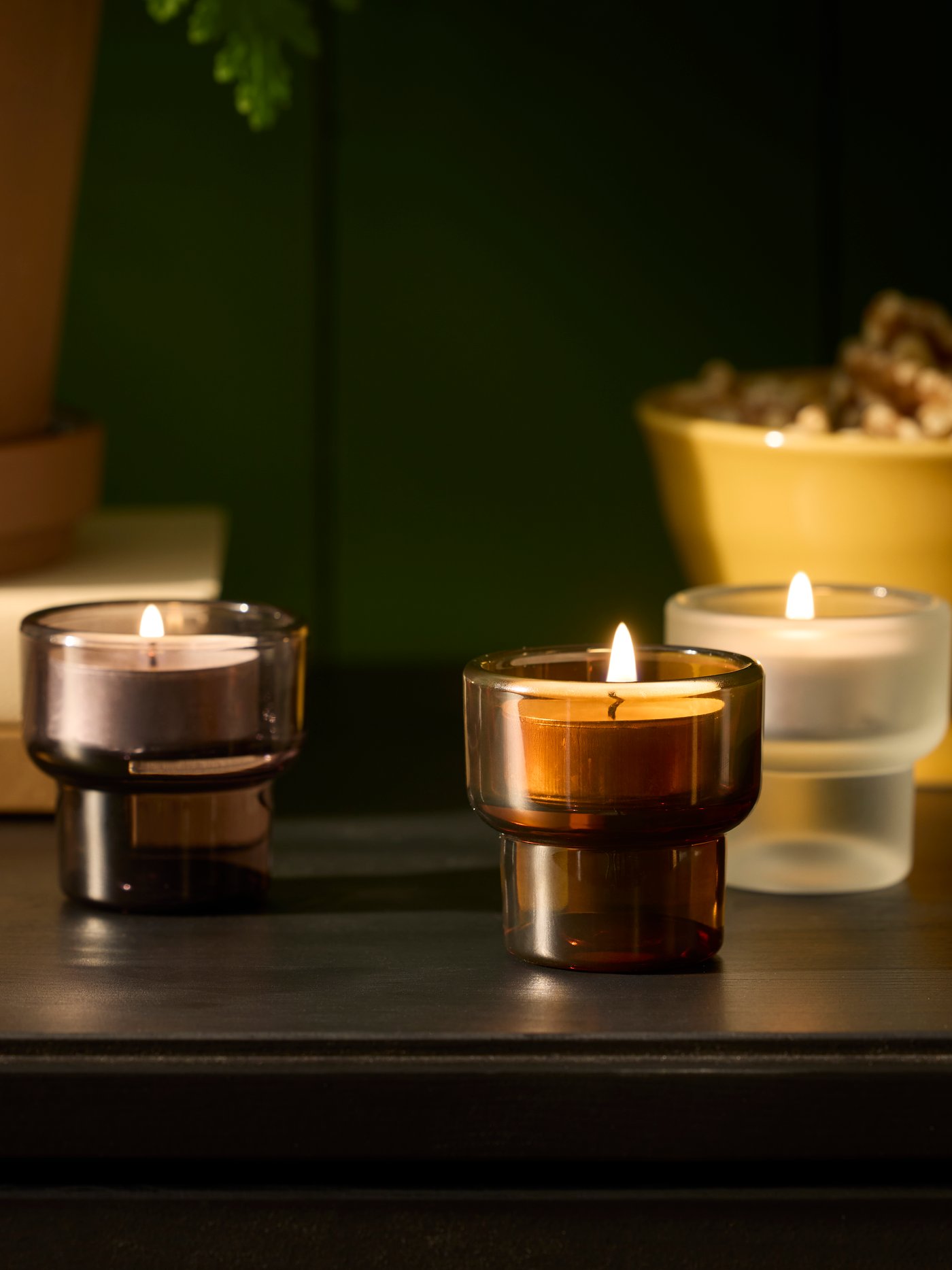 Three LADUSVALA tealight holders in mixed colours are arranged on a dark table, together with a plant and a decorative bowl.
