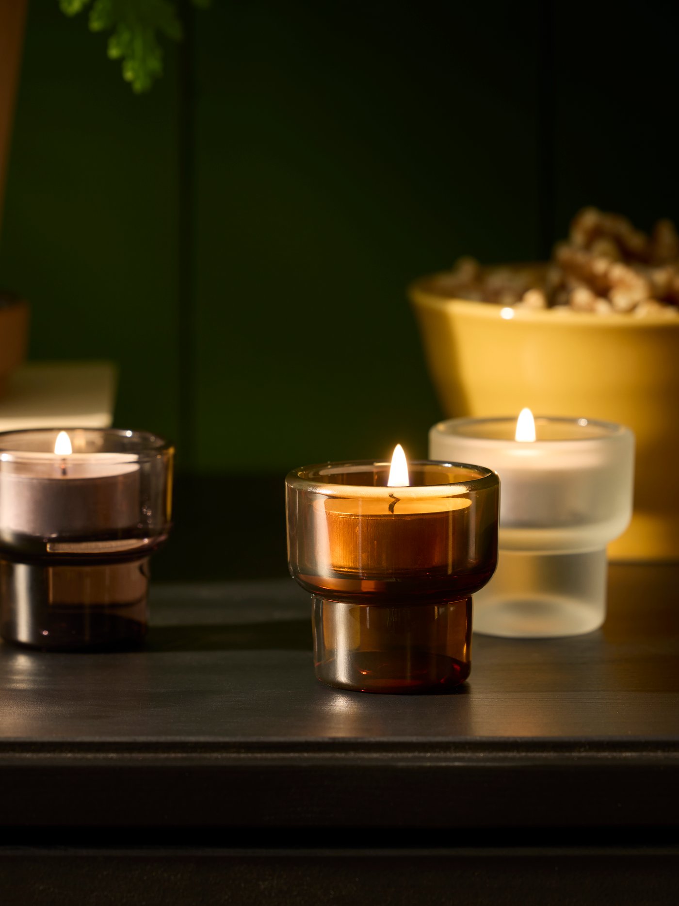 Three LADUSVALA tealight holders in mixed colours are arranged on a dark table, together with a plant and a decorative bowl.