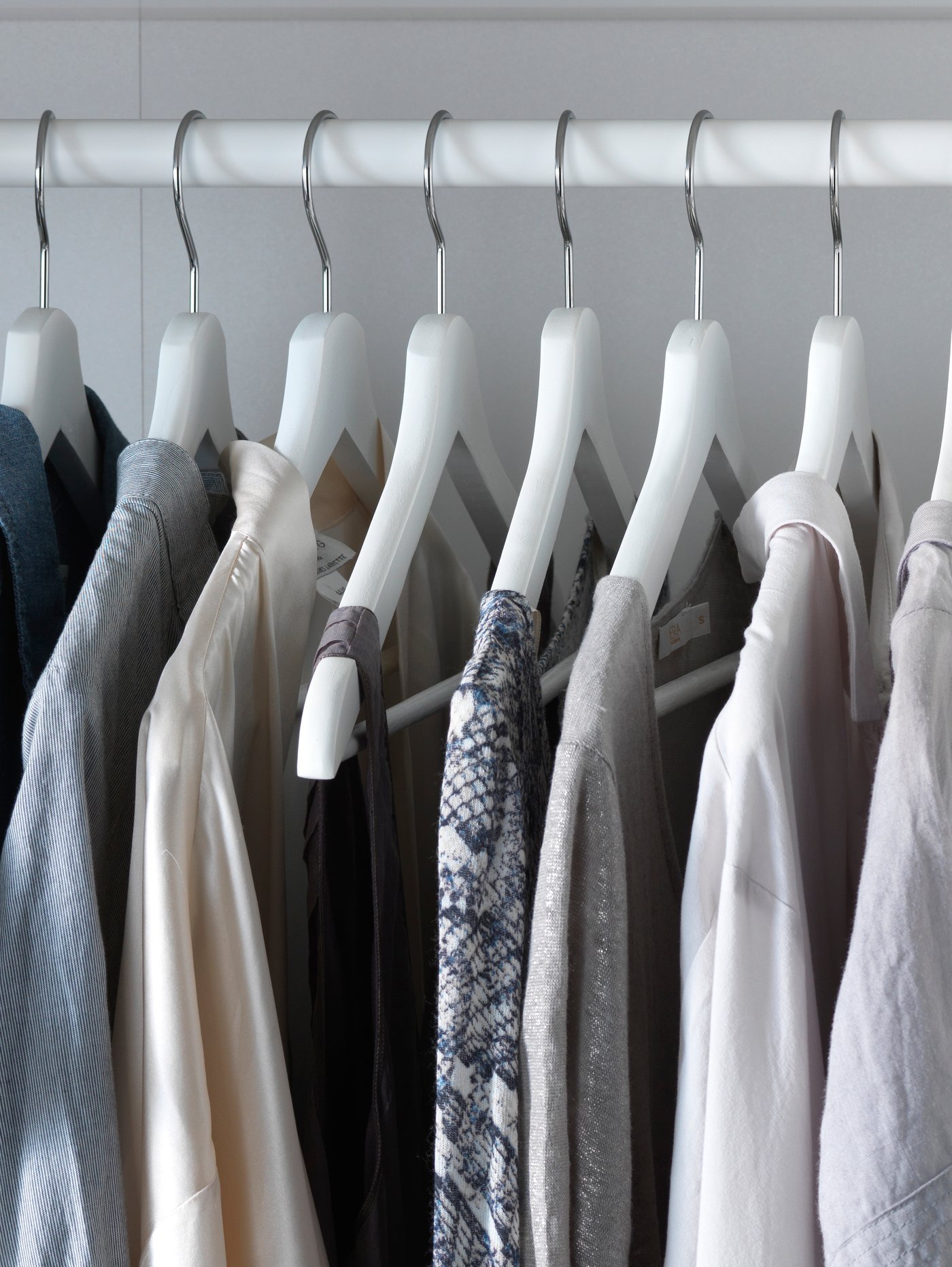 Detalle de siete perchas de plástico blancas en una barra para colgar la ropa con varias prendas de mujer.