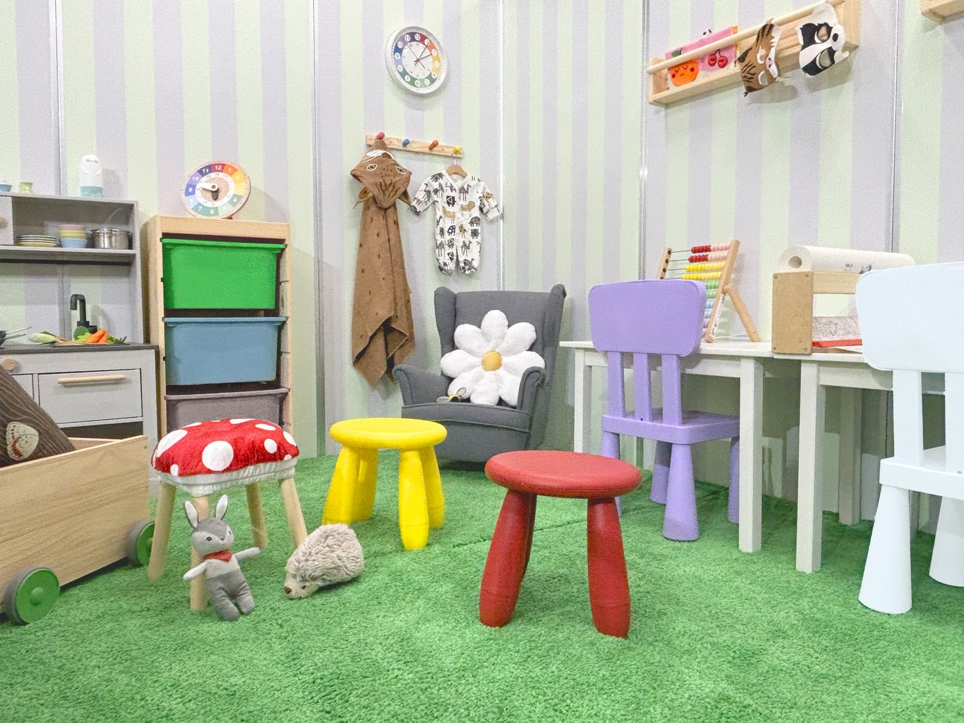 緑のカーペットとストライプ柄の壁紙が特徴の、カラフルな子供部屋。デイジーのクッションが置かれた小さなアームチェア、カラフルなスツール、子供用のテーブル、おもちゃのキッチン、そしてそろばんやぬいぐるみといった様々なおもちゃが置かれています。