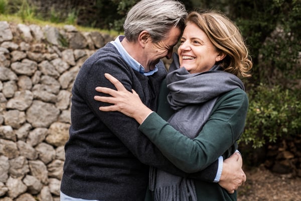 Un homme aux cheveux gris enlace son épouse à l’extérieur. Ils sourient tous les deux. 