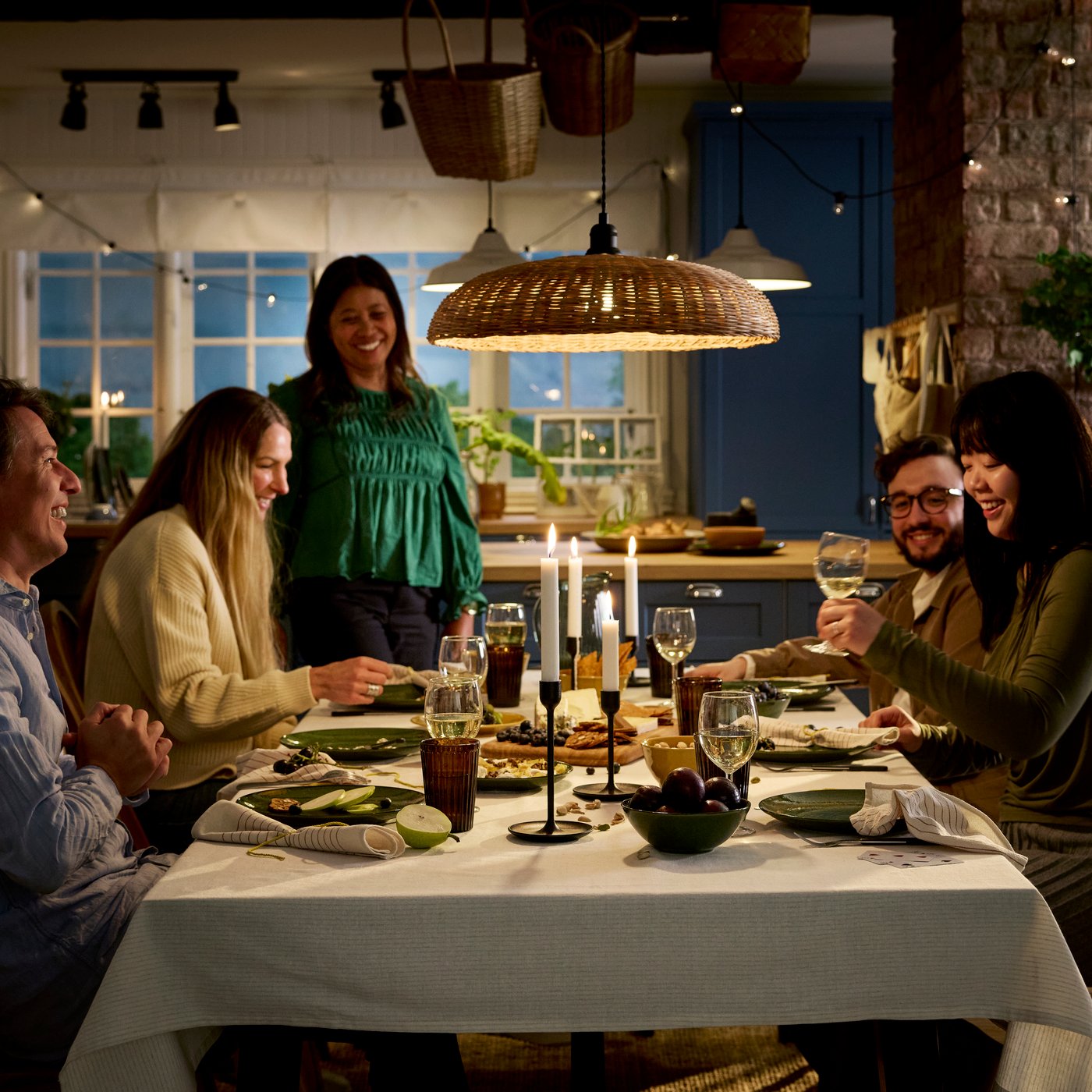 People are gathered around a dinner table for a meal of cheese and fresh fruit, all in a cosy, atmosphere with candles.