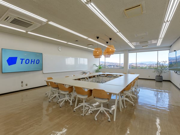大きな窓から光が入る会議スペース。長い木製テーブルの周りに白いチェアが並び、前方のモニターにロゴが表示されている。