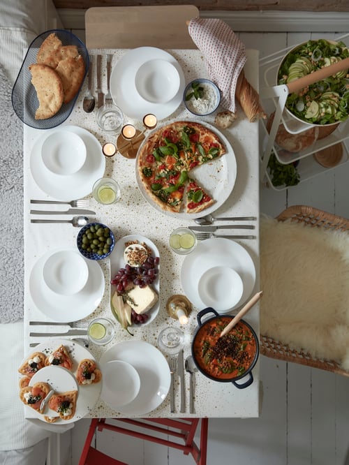A white table set with lots of plates, cutlery and food, and an OFTAST plate in white with a large pizza on it.