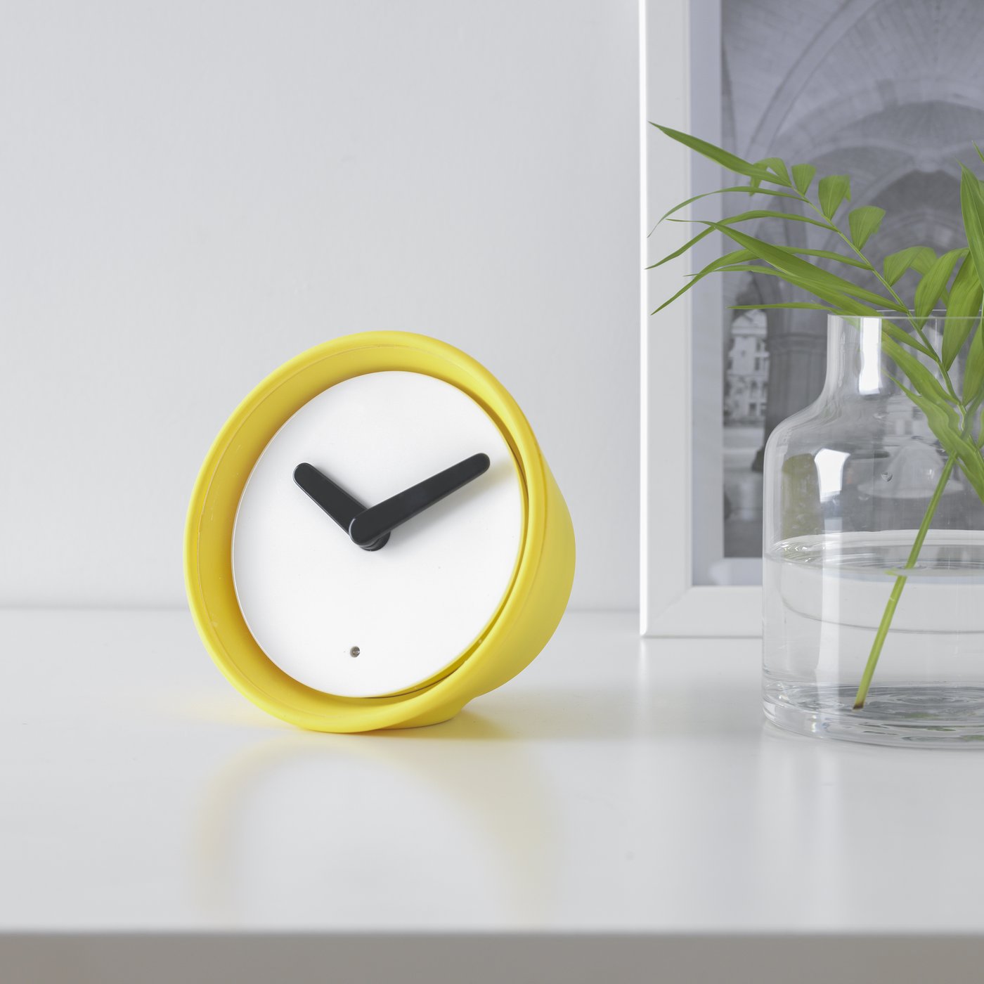 Reloj amarillo sobre una cubierta blanca junto a un florero de vidrio transparente.
