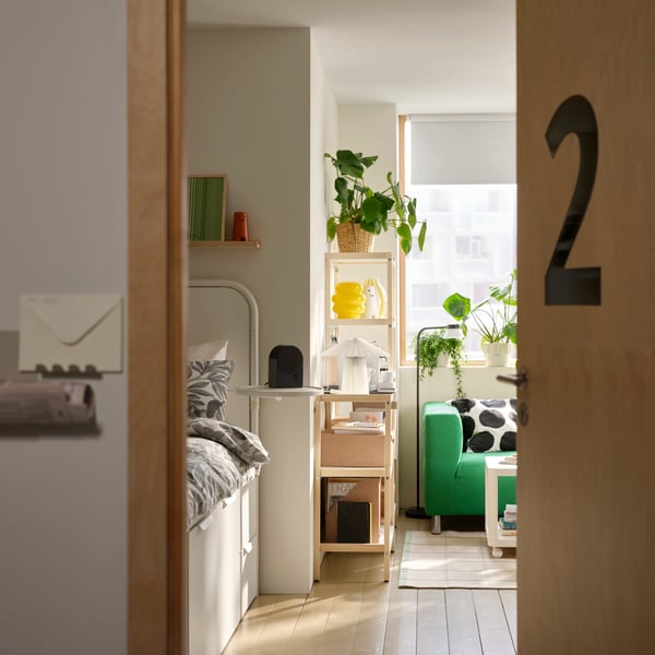 A modern student room rental has its door open, showing parts of EKENABBEN shelving units, a white bed and a green sofa.