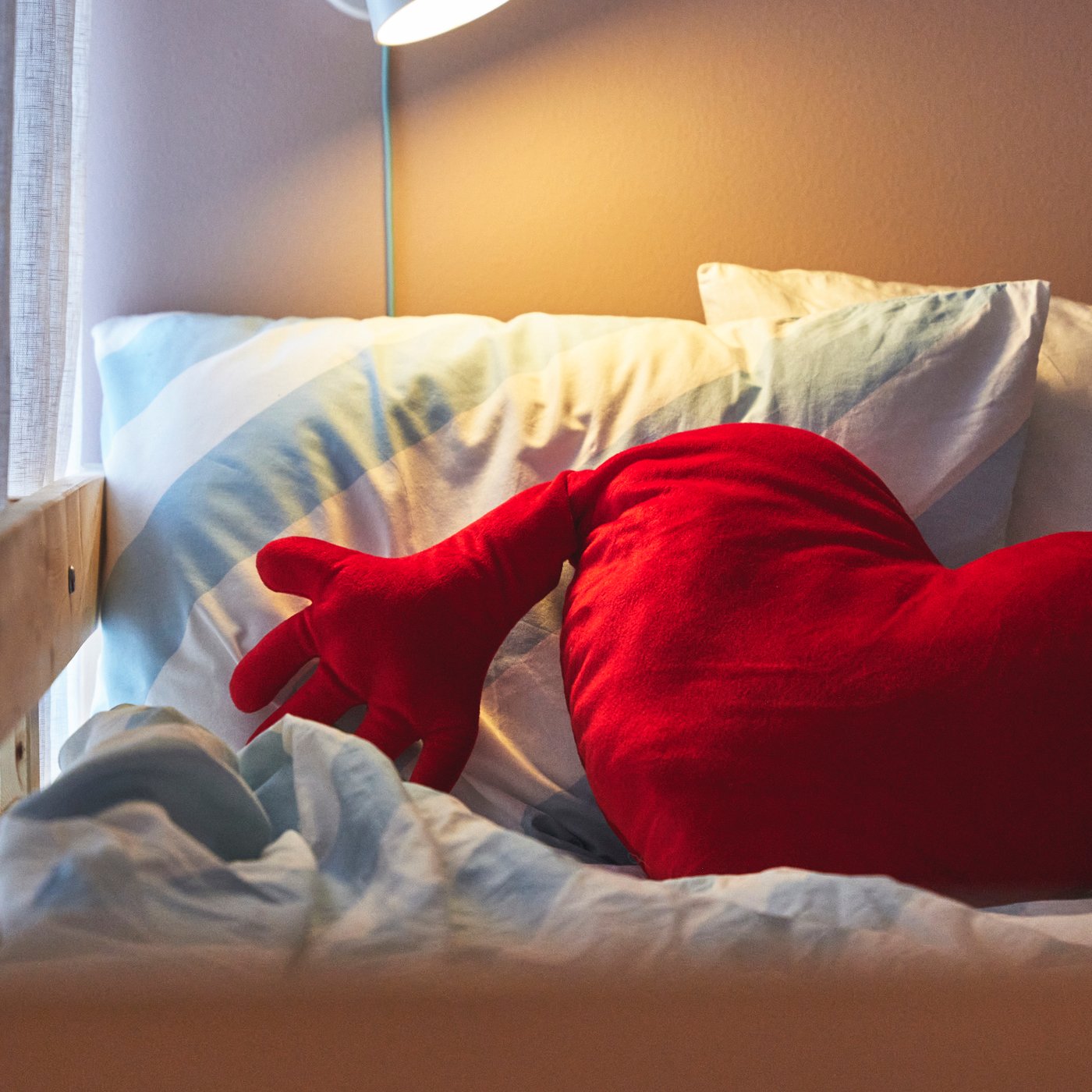 En röd hjärtformad kudde med armar ligger på en bäddad säng med ljusa täcken, upplyst av en lampa vid huvudändan.