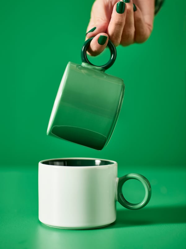 A person with green-coloured nails is holding a green PIGGÅL cup just over a white PIGGÅL mug.