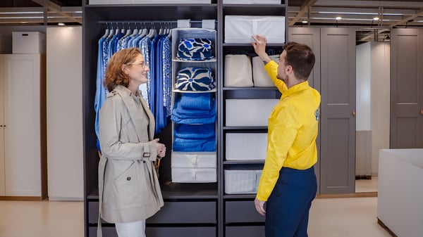 Konsultant IKEA stojący w żółtej koszulce przy szafie PAX w sklepie IKEA i pokazujący rozwiązania do przechowywania. Przy szafie stoi również klientka, która omawia z konsultantem dostępne rozwiązania.