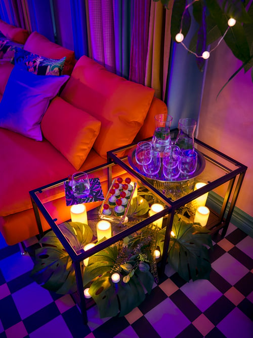 Des tables gigognes VITTSJÖ brun noir/verre sont placées l’une sur l’autre, avec des boissons sur le plateau et un décor luxuriant sur la tablette inférieure.