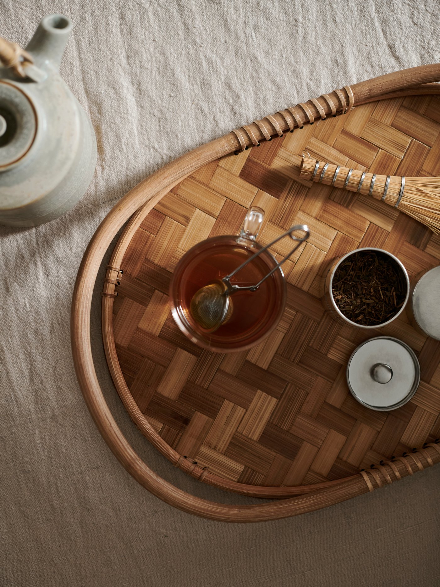 Nahaufnahme eines geflochtenen Tabletts mit einer Tasse Tee, losem Tee, einem Schneebesen und Keramikbehältern auf einem strukturierten Tuch.