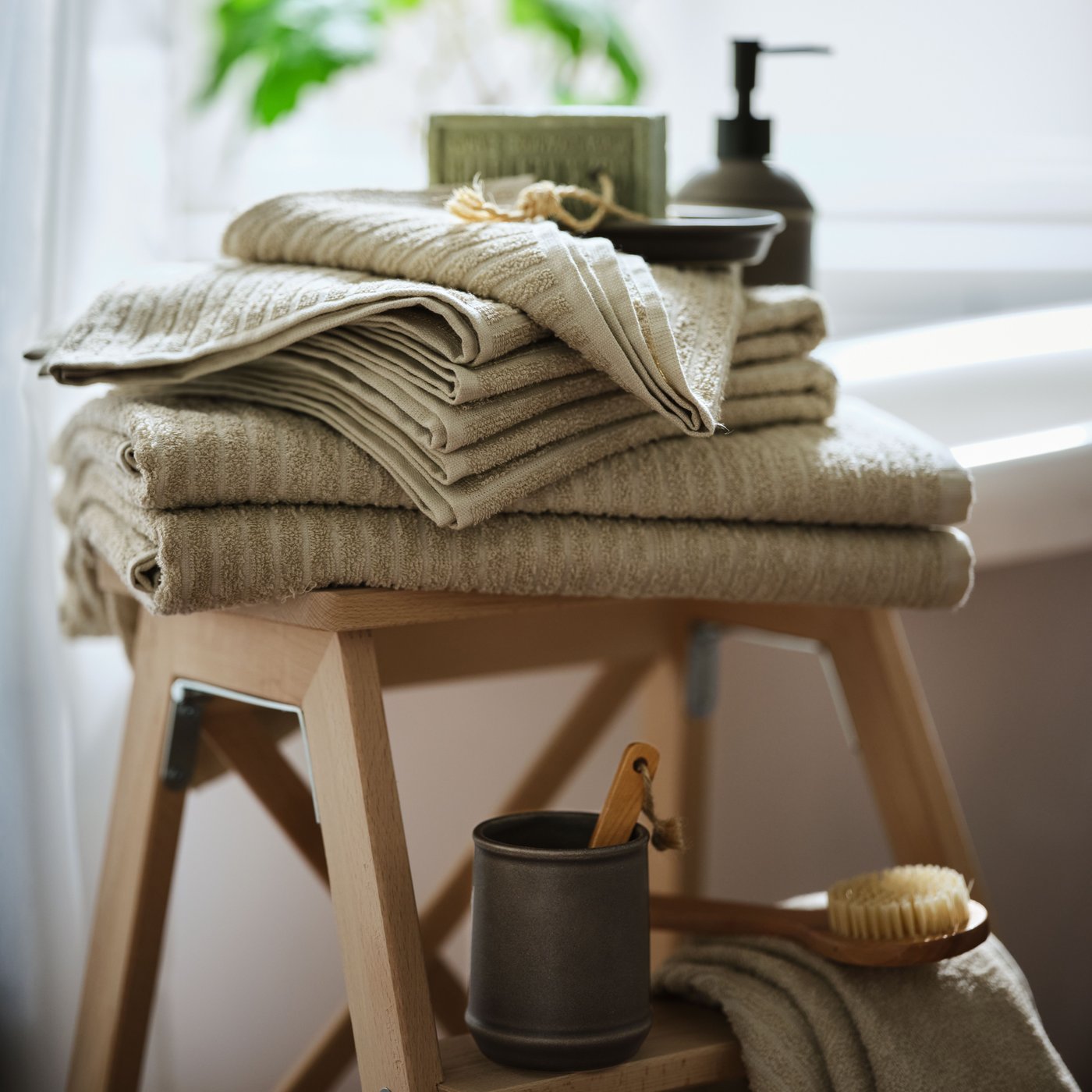 A pile of VÅGSJÖN cotton towels in beige colour on a wooden stool in front of a window. Soap and a body brush next to it.  