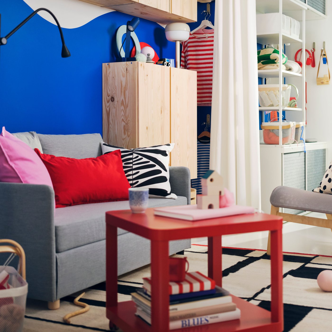 A light, brightly coloured living room with a light-grey FRIDHULT sofa-bed, a red TINGBY side table and plenty of textiles.