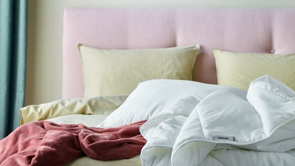 Close-up of a light pink upholstered bed with bedding in light yellow and dark pink. A white duvet and pillow on top.
