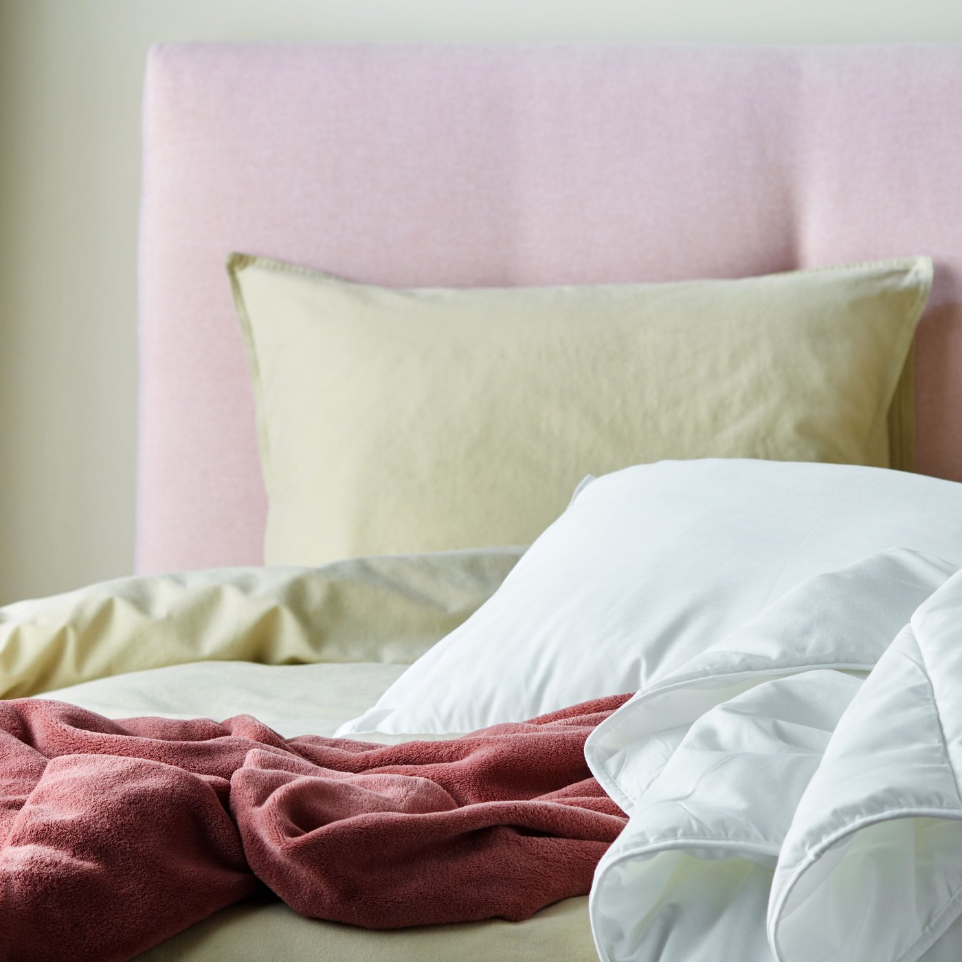 Close-up of a light pink upholstered bed with bedding in light yellow and dark pink. A white duvet and pillow on top.