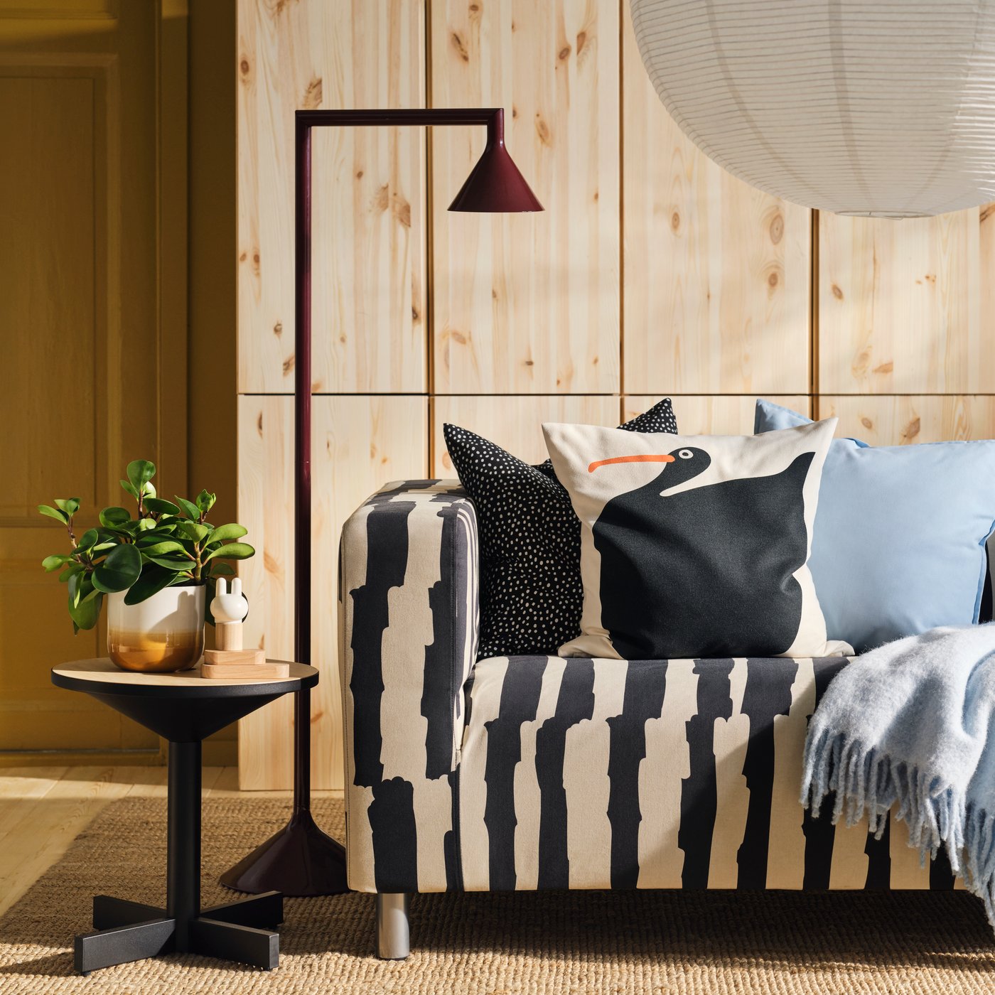 A black/beige striped KLIPPAN sofa in a living room with a wooden wall, a side table and a burgundy floor lamp.
