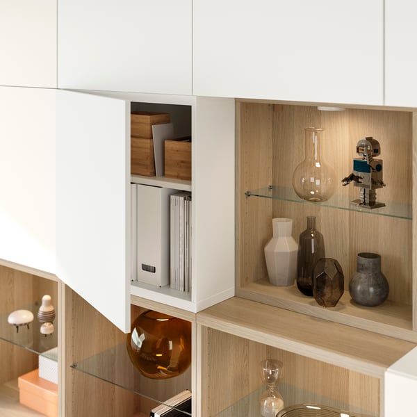 A close-up of a white stained oak/white LAPPVIKEN BESTÅ storage combination shows various objects displayed on glass shelves.