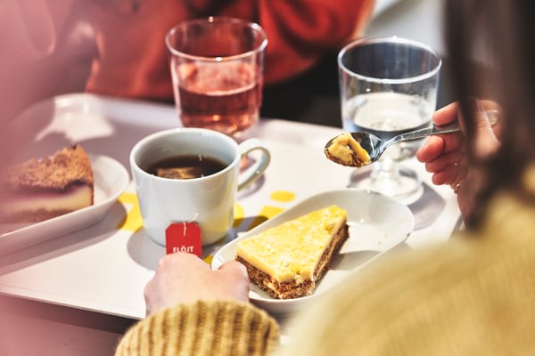 A person wearing a cosy jumper is enjoying a slice of cake on a tray, accompanied by tea and water. The warm colours create a relaxed and inviting atmosphere.