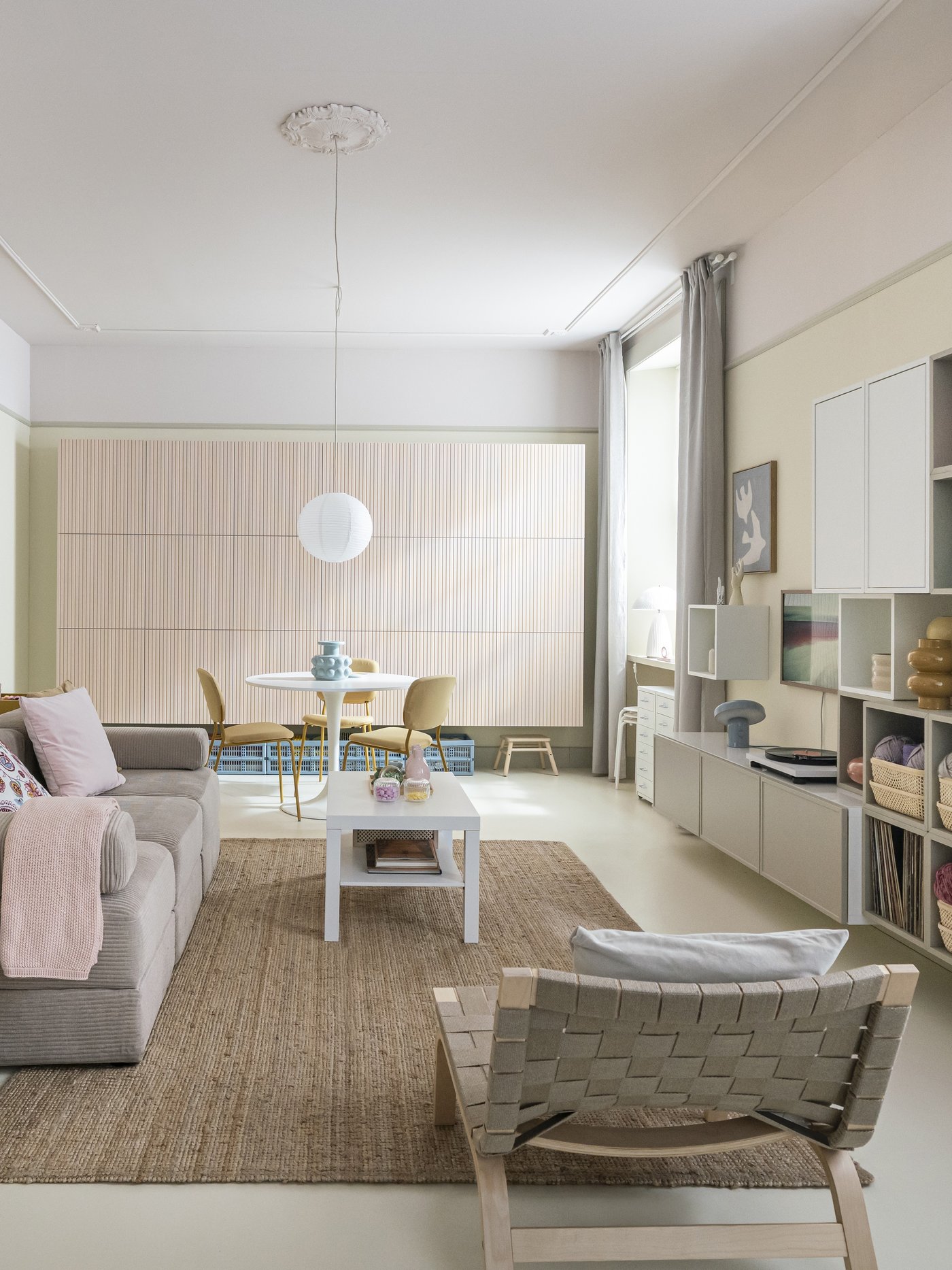 Soggiorno luminoso arredato con mobili IKEA. È presente undivano grigio chiaro, un tappeto naturale, degli scaffali bianchi e una zona pranzo sullo sfondo.