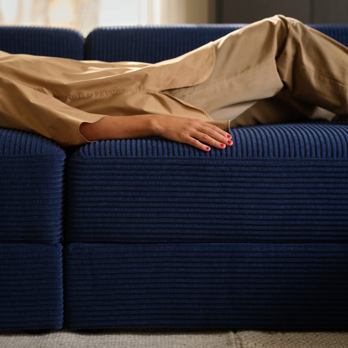 A person lies down flat on a dark-blue JÄTTEBO four-seat modular sofa, in turn placed in a softly sunlit living room.