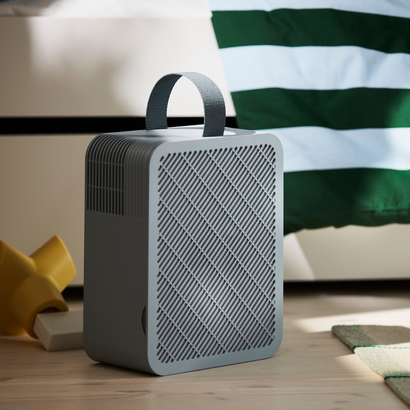 A light blue UPPÅTVIND air purifier on a wooden floor in a child’s room next to a white bed with a striped duvet cover.