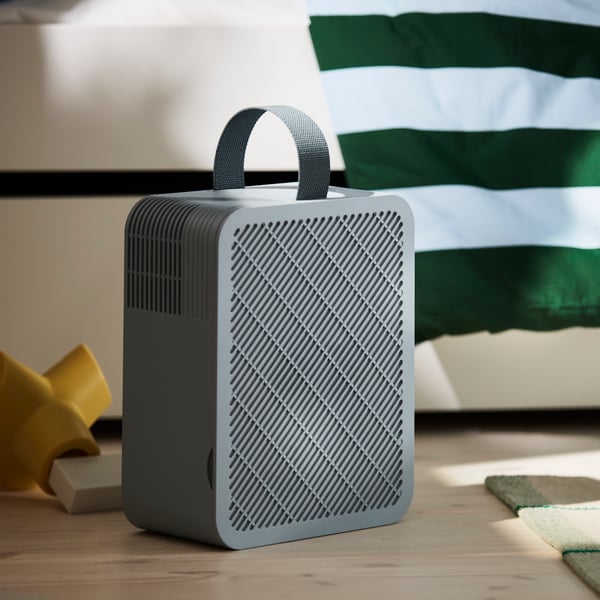 A light blue UPPÅTVIND air purifier on a wooden floor in a child’s room next to a white bed with a striped duvet cover.