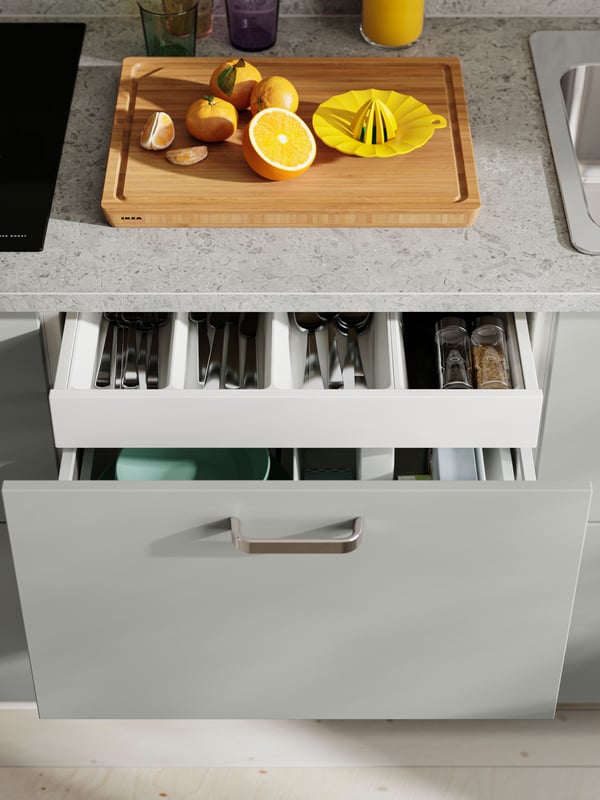 A drawer with HAVSTORP light grey fronts with organized utensils, with a cutting board and a citrus juicer on the countertop.