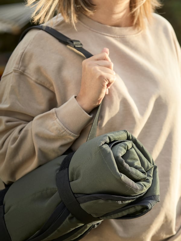 The FJÄLLMOTT picnic blanket is folded up neatly and is being carried using its convenient straps on a woman's shoulder.
