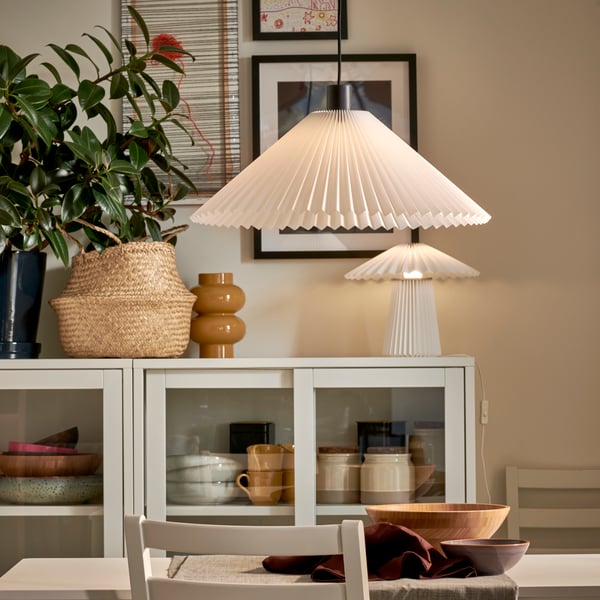 A dining area with a light colour scheme, glass-door cabinets, plants and art, all lit by a KUDDLAVA pendant and table lamp.