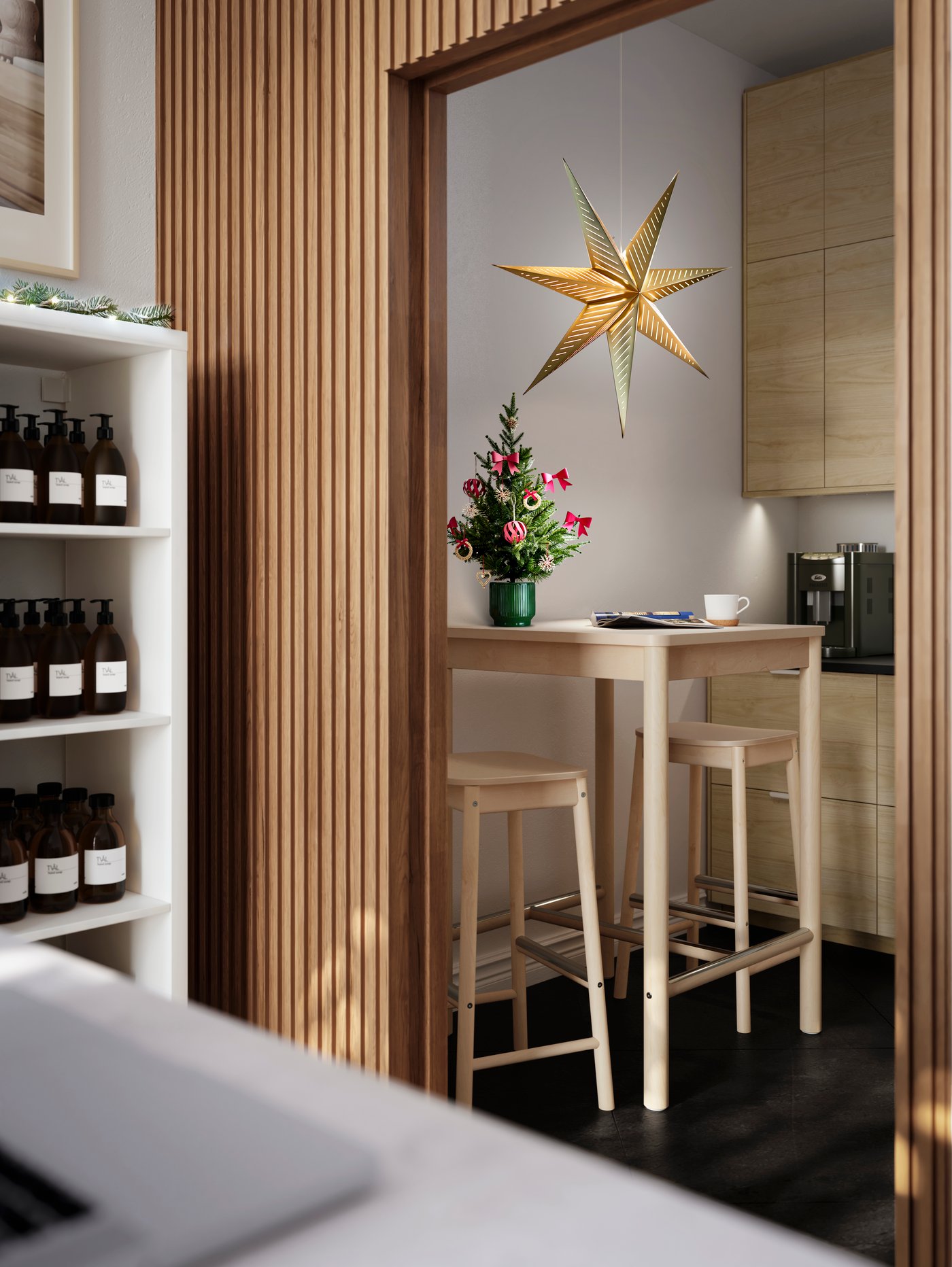 Un'apertura nella parete in legno listellare da cui si accede a una stanza sul retro dove ci sono un tavolo con sgabelli bar in betulla RÖNNINGE, alberello di Natale decorato con VINTERFINT e una lampada STRÅLA.