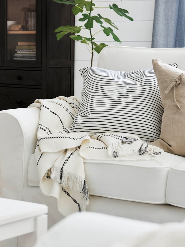 Un coussin enveloppé dans une housse INGALILL blanche avec des rayures gris foncé et un coussin avec une housse AINA beige sont posés sur un canapé UPPLAND blanc.