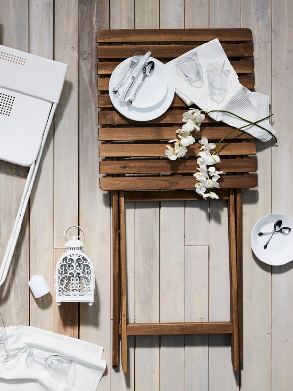 Un tavolo da giardino ASKHOLMEN piegato con sopra un piatto, delle posate e due bicchieri su un tovagliolo.