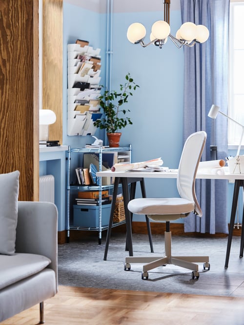 Bright office with beige office chair with steel frame and mesh backrest, white desk, beige table lamp, and 7-arm chandelier.