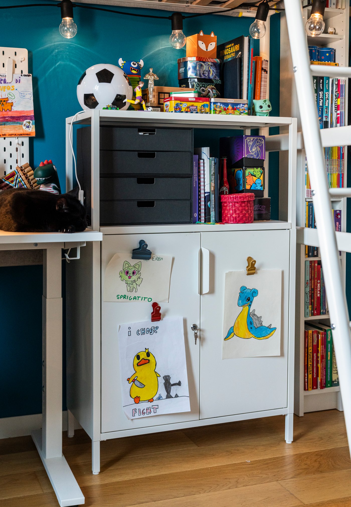 Mobile bianco in cameretta decorato con disegni di bambini sulle ante, scaffali superiori pieni di giochi, libri e scatole portaoggetti.