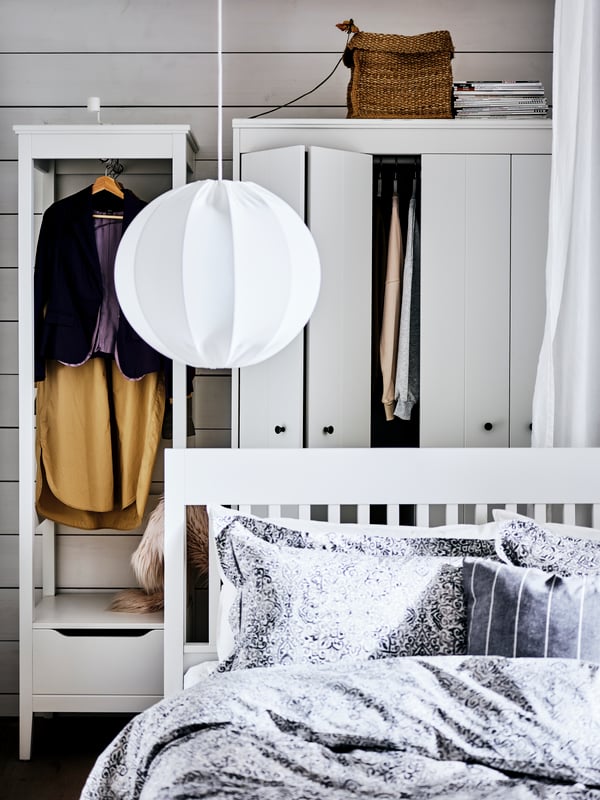 A white IDANÄS bedframe with a pendant lamp above it, in front of two white wardrobes with hanging clothes on show.